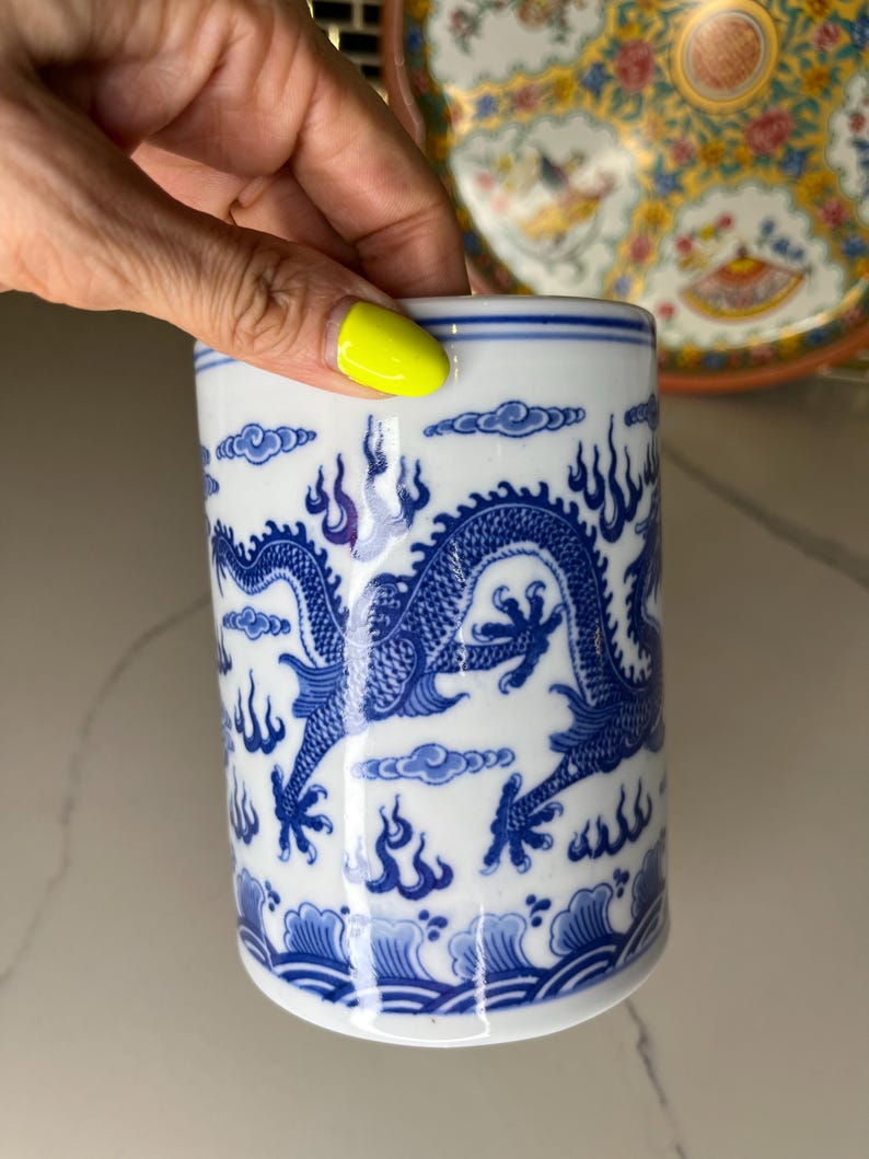 May include: A white and blue ceramic cup with a dragon design. The dragon is blue and wraps around the cup, with flames and clouds. The cup has a blue rim and base. A hand with yellow nail polish is holding the cup.