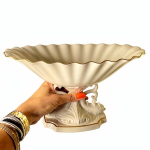 May include: A cream-colored ceramic pedestal bowl featuring a wide, scalloped rim with a thin gold-painted edge. The base of the bowl is supported by an ornate, sculpted stand with intricate relief detailing and a matching gold-painted trim.
