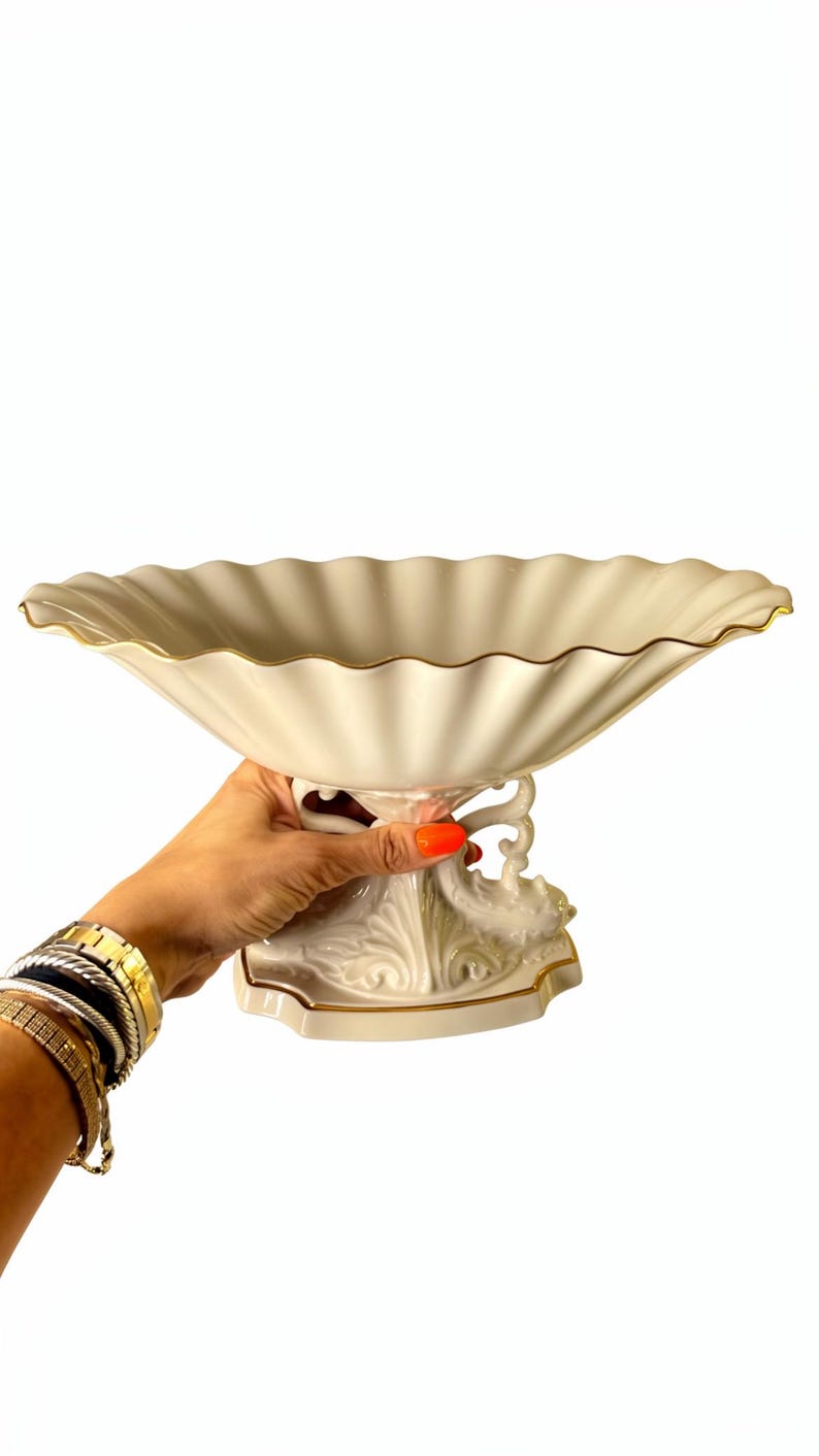 May include: A cream-colored ceramic pedestal bowl featuring a wide, scalloped rim with a thin gold-painted edge. The base of the bowl is supported by an ornate, sculpted stand with intricate relief detailing and a matching gold-painted trim.