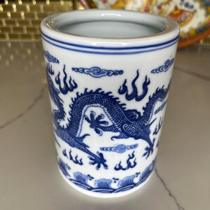 May include: A white ceramic cup with a blue dragon design. The cup has a cylindrical shape with a blue rim and a detailed dragon illustration wrapping around the body. The dragon is depicted with intricate scales and flowing lines.