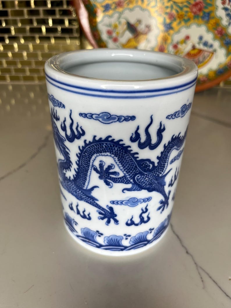 May include: A white ceramic cup with a blue dragon design. The cup has a cylindrical shape with a blue rim and a detailed dragon illustration wrapping around the body. The dragon is depicted with intricate scales and flowing lines.