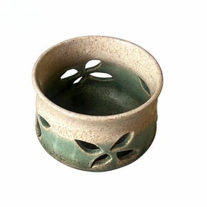 Puede incluir: Un cuenco de cerámica con una sección superior de color crema moteado y una sección inferior de color verde salvia. El cuenco está decorado con formas de mariposa recortadas en los lados, revelando el interior con un esmalte verde oscuro.