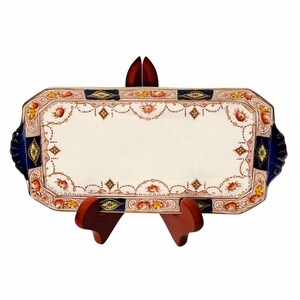 Puede incluir: Una bandeja de servicio rectangular de cerámica con un centro color crema y un borde decorativo con patrones florales en naranja, amarillo y verde. Los bordes están acentuados con paneles azul oscuro y ribetes dorados, con asas festoneadas.