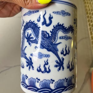 May include: White ceramic cylindrical container with a blue dragon design. The container features a detailed illustration of a dragon, clouds, and flames. The base has a wave pattern. The container is held by a hand.