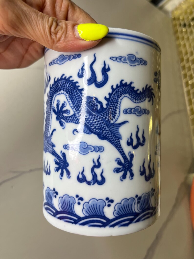 May include: White ceramic cylindrical container with a blue dragon design. The container features a detailed illustration of a dragon, clouds, and flames. The base has a wave pattern. The container is held by a hand.