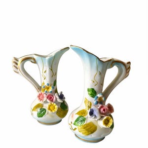 Peut inclure: Deux vases en céramique blanche avec un bord et une anse bleu clair, ornés de compositions florales colorées. Les vases ont une bordure dorée et mesurent environ 15 cm de haut.