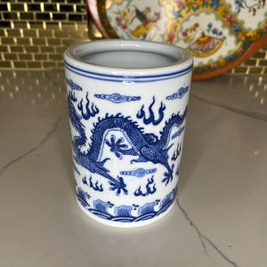 May include: A cylindrical ceramic container with a blue and white dragon design. The container has a white interior and a blue rim. The dragon is depicted with intricate details, surrounded by stylized clouds and flames. The container is approximately 4 inches tall.