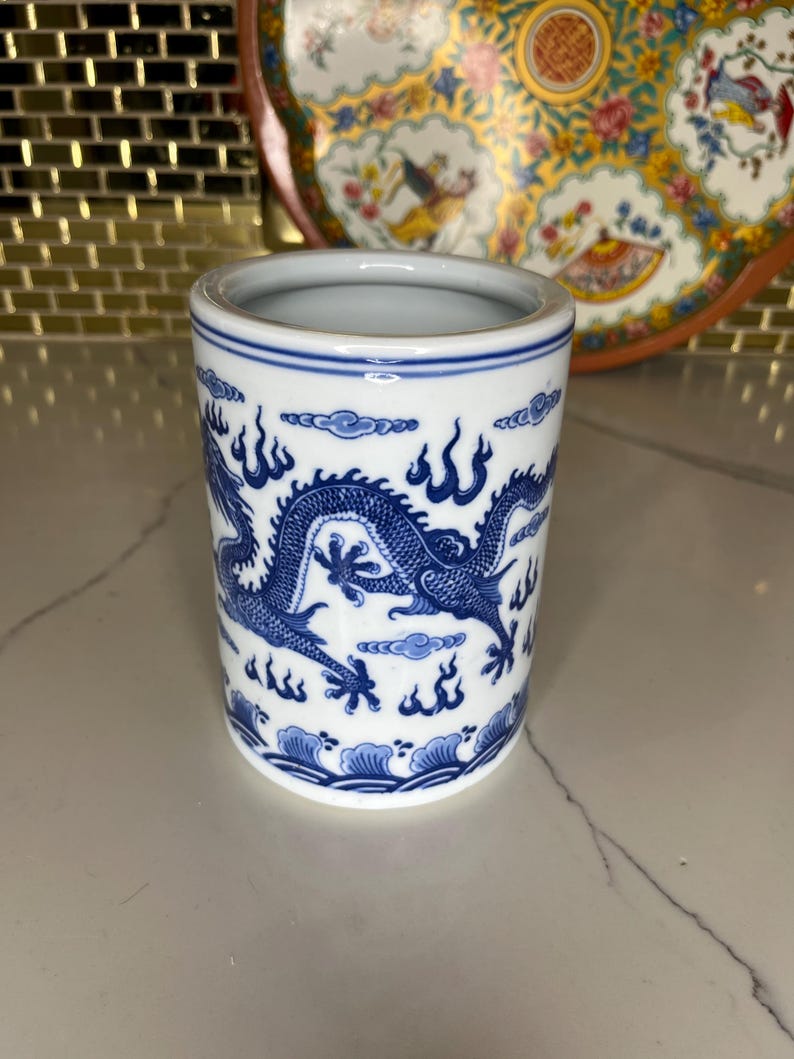 May include: A cylindrical ceramic container with a blue and white dragon design. The container has a white interior and a blue rim. The dragon is depicted with intricate details, surrounded by stylized clouds and flames. The container is approximately 4 inches tall.