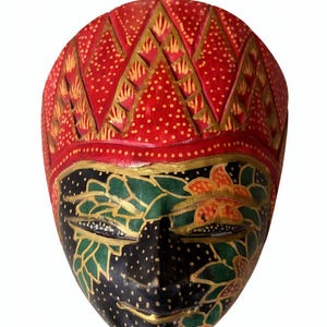 May include: A hand-carved wooden mask with a red, gold, black, and green colour scheme. The mask features a red headdress with gold accents and a black face adorned with green and gold floral patterns. The mask is a decorative item.