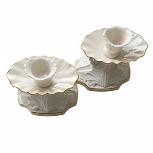 May include: Two white ceramic candle holders featuring a scalloped, flower-like top edge and a base with embossed leaf-like patterns. Each holder is finished with a thin gold trim along the edges of the top plate and the base perimeter.