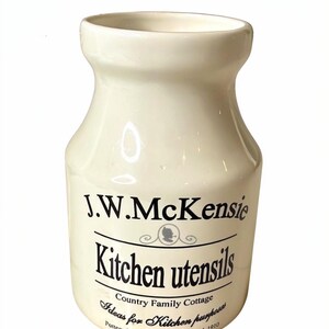 May include: Cream-coloured ceramic kitchen utensil holder with a wide opening and a slightly tapered body. The holder features the text "J.W. McKensie Kitchen utensils" and "Country Family Cottage" with additional text below. A vintage kitchen accessory.