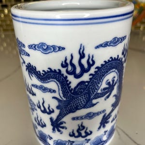 May include: White ceramic tumbler with a blue dragon design. The tumbler features a detailed illustration of a dragon, clouds, and flames. The rim and base have blue accents. A decorative piece for the home.