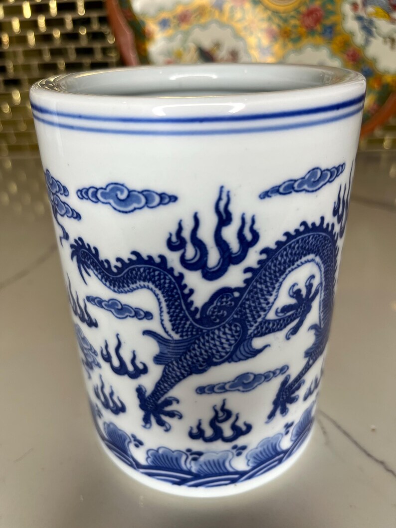 May include: White ceramic tumbler with a blue dragon design. The tumbler features a detailed illustration of a dragon, clouds, and flames. The rim and base have blue accents. A decorative piece for the home.