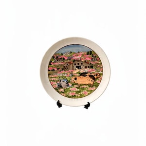 May include: A round ceramic plate featuring a colorful illustration of a farm scene with two spotted pigs in the foreground, a wooden barn, rolling green hills, pink flowering bushes, and a blue sky, all set against a plain white background.