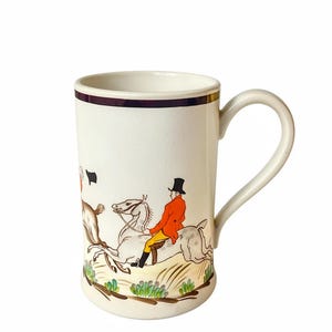 May include: Cream-colored ceramic mug with a hunting scene illustration. The design features a rider on horseback in a red coat, a dog, and a black flag. The mug has a dark purple band near the rim and a curved handle.
