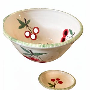 Peut inclure: Ensemble de bol et de petite assiette en céramique crème. Le bol et l'assiette présentent un motif de cerises peint à la main avec des cerises rouges et des feuilles vertes. Les bords ont une bordure verte texturée.