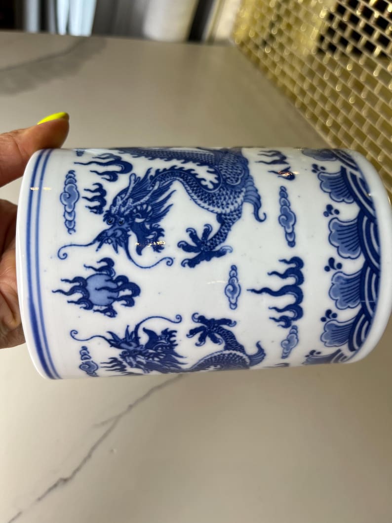 May include: White ceramic cylinder with blue dragon and cloud designs. The detailed artwork features a traditional Asian aesthetic. The cylindrical shape is accented with a blue border at the top and bottom. The design is repeated around the entire cylinder.