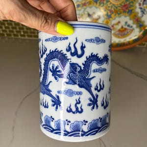 May include: A cylindrical white ceramic container with a blue dragon design. The dragon is depicted with intricate details, surrounded by stylized clouds and flames. The container has a wave pattern at the base and a blue band at the top.