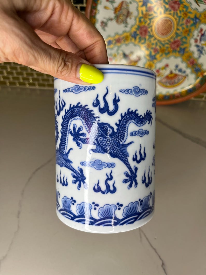 May include: A cylindrical white ceramic container with a blue dragon design. The dragon is depicted with intricate details, surrounded by stylized clouds and flames. The container has a wave pattern at the base and a blue band at the top.