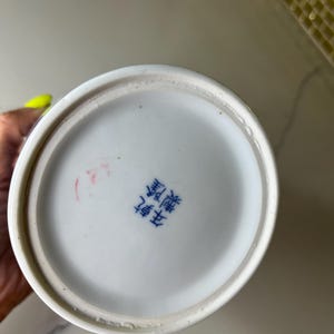 May include: A white ceramic vase base with a blue rim and a Chinese character stamp. The base is circular and has a slightly rough texture. The stamp is in blue ink and appears to be a maker's mark.