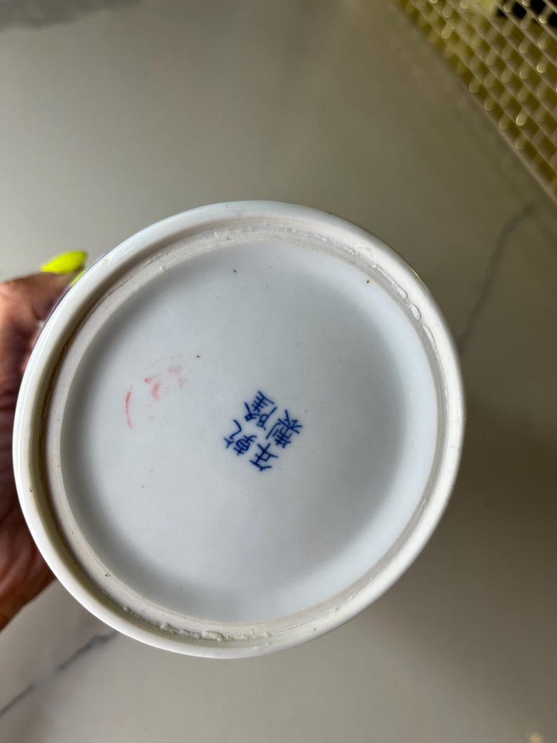 May include: A white ceramic vase base with a blue rim and a Chinese character stamp. The base is circular and has a slightly rough texture. The stamp is in blue ink and appears to be a maker's mark.