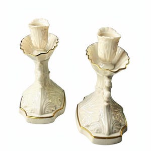 May include: A pair of off-white porcelain candle holders featuring intricate embossed leaf patterns, scalloped edges, and delicate gold trim along the rims and base. Each holder has a central cup for a candle and a wide, ornate pedestal base.