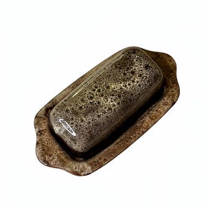 May include: A brown butter dish with a lid, featuring a speckled pattern. The dish has a rectangular shape with a slightly raised edge. The lid is rectangular with rounded edges and a glossy finish.