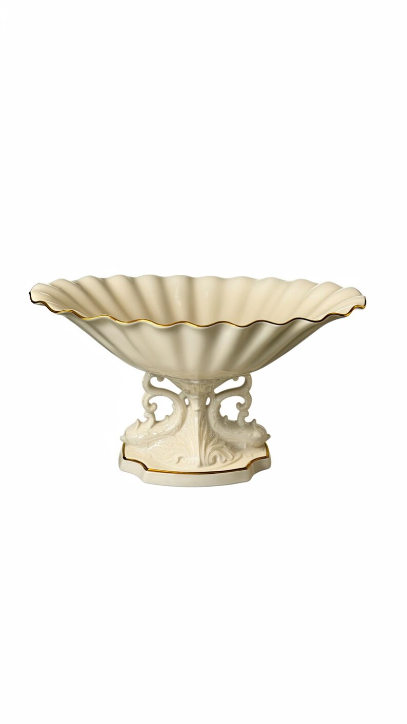 May include: A cream-colored ceramic pedestal bowl with a scalloped rim and a gold-trimmed edge. The base features intricate sculpted dolphin motifs and a matching gold-trimmed foot, providing a decorative support for the wide, fluted basin.
