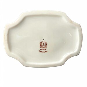 May include: The underside of a white porcelain object with a scalloped edge. Centered is a brown stamp featuring a wreath, the letter L, the word LENOX, and the text MADE IN USA. The surface is smooth and glossy with a small pinhole near the mark.