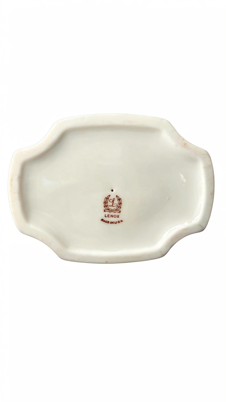 May include: The underside of a white porcelain object with a scalloped edge. Centered is a brown stamp featuring a wreath, the letter L, the word LENOX, and the text MADE IN USA. The surface is smooth and glossy with a small pinhole near the mark.