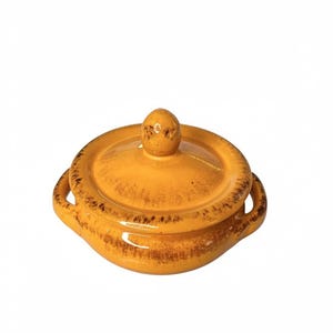 Puede incluir: Un cuenco de cerámica con tapa de color amarillo cálido, con forma redondeada, dos asas y tapa a juego. El cuenco tiene un aspecto texturizado y rústico con detalles en marrón oscuro en los bordes.