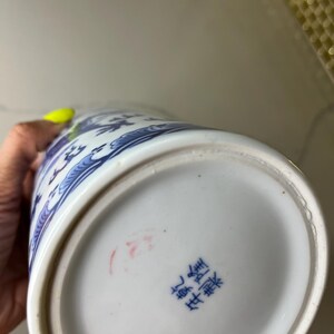 May include: A white and blue ceramic vase with a detailed floral design. The vase has a rounded base and a slightly flared top. The bottom of the vase has blue Chinese characters and a red stamp.