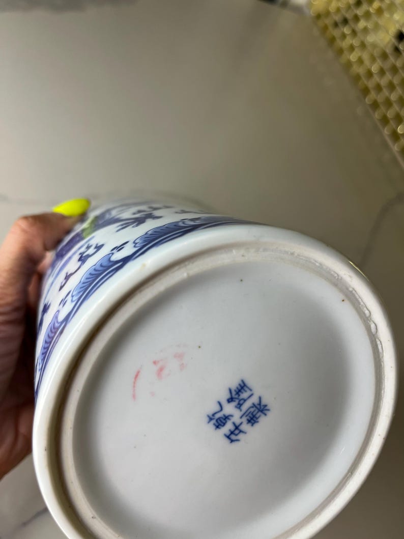 May include: A white and blue ceramic vase with a detailed floral design. The vase has a rounded base and a slightly flared top. The bottom of the vase has blue Chinese characters and a red stamp.