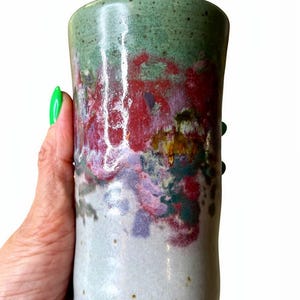 May include: A handmade ceramic tumbler with a cylindrical shape. The top is a mottled green, transitioning to a vibrant mix of red, purple, and yellow hues in the middle. The base is a light, speckled colour.