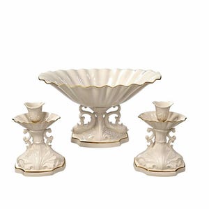 May include: A set of three off-white ceramic pieces featuring a large scalloped bowl and two matching candle holders. Each piece has an ornate, sculpted base with intricate scrollwork details and a thin gold trim along the edges.
