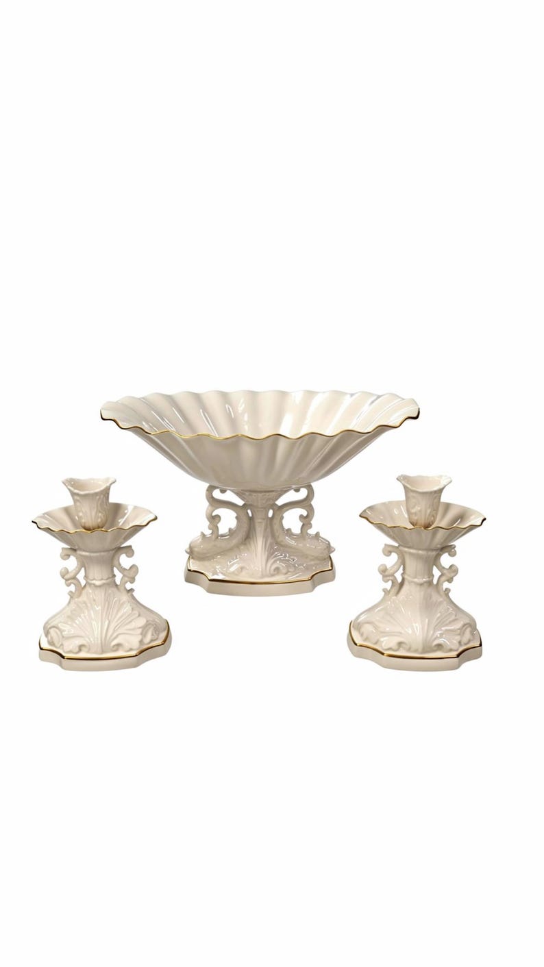 May include: A set of three off-white ceramic pieces featuring a large scalloped bowl and two matching candle holders. Each piece has an ornate, sculpted base with intricate scrollwork details and a thin gold trim along the edges.