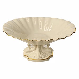 May include: A cream-colored ceramic pedestal bowl with a wide, scalloped rim and a gold-painted edge. The base features sculpted swan figures on either side, also accented with gold trim, supporting the shallow, fluted basin of the dish.