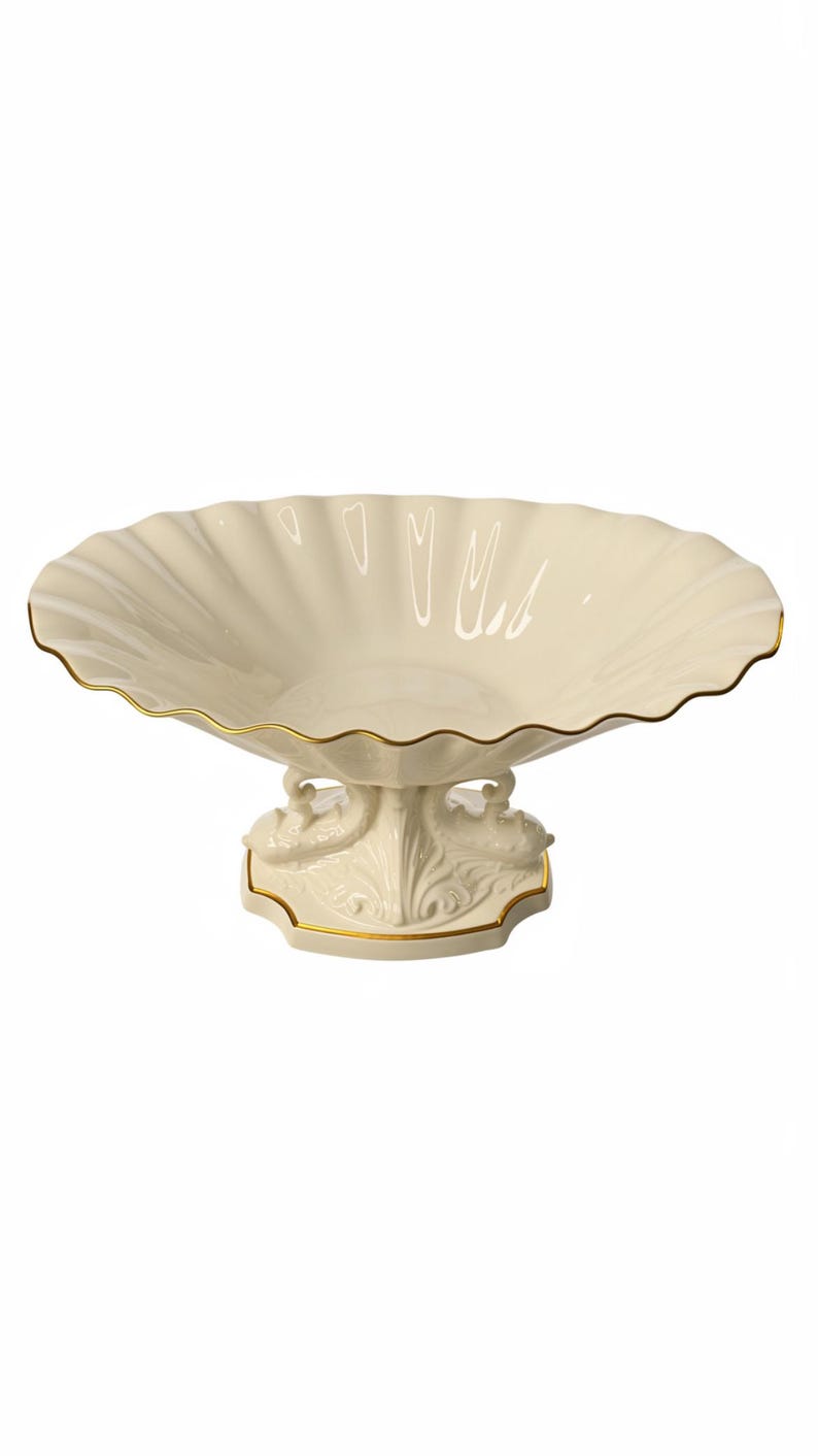 May include: A cream-colored ceramic pedestal bowl with a wide, scalloped rim and a gold-painted edge. The base features sculpted swan figures on either side, also accented with gold trim, supporting the shallow, fluted basin of the dish.