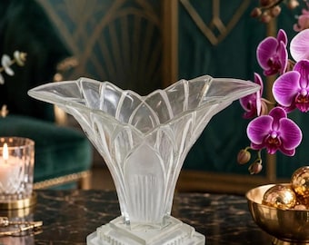 May include: A clear glass vase with a wide, flared rim and a geometric, petal-like structure. The vase features a square, frosted glass base with vertical ribbed detailing and a smooth, transparent upper section that tapers toward the center.