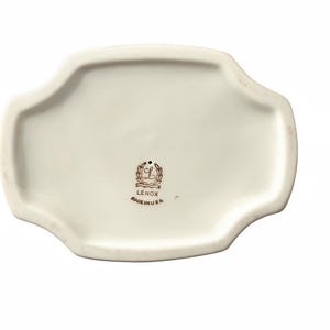 May include: An off-white ceramic tray with a scalloped edge and a smooth, glossy finish. The center features a black stamp with a laurel wreath, the letter L, the text LENOX, and the text MADE IN USA, indicating the origin of this ceramic piece.