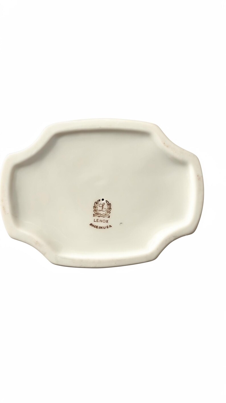 May include: An off-white ceramic tray with a scalloped edge and a smooth, glossy finish. The center features a black stamp with a laurel wreath, the letter L, the text LENOX, and the text MADE IN USA, indicating the origin of this ceramic piece.