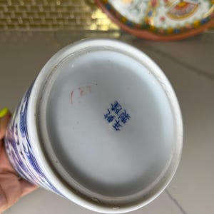 May include: A white and blue porcelain vase with a floral design. The bottom of the vase has blue Chinese characters. A decorative plate with a floral design is in the background.