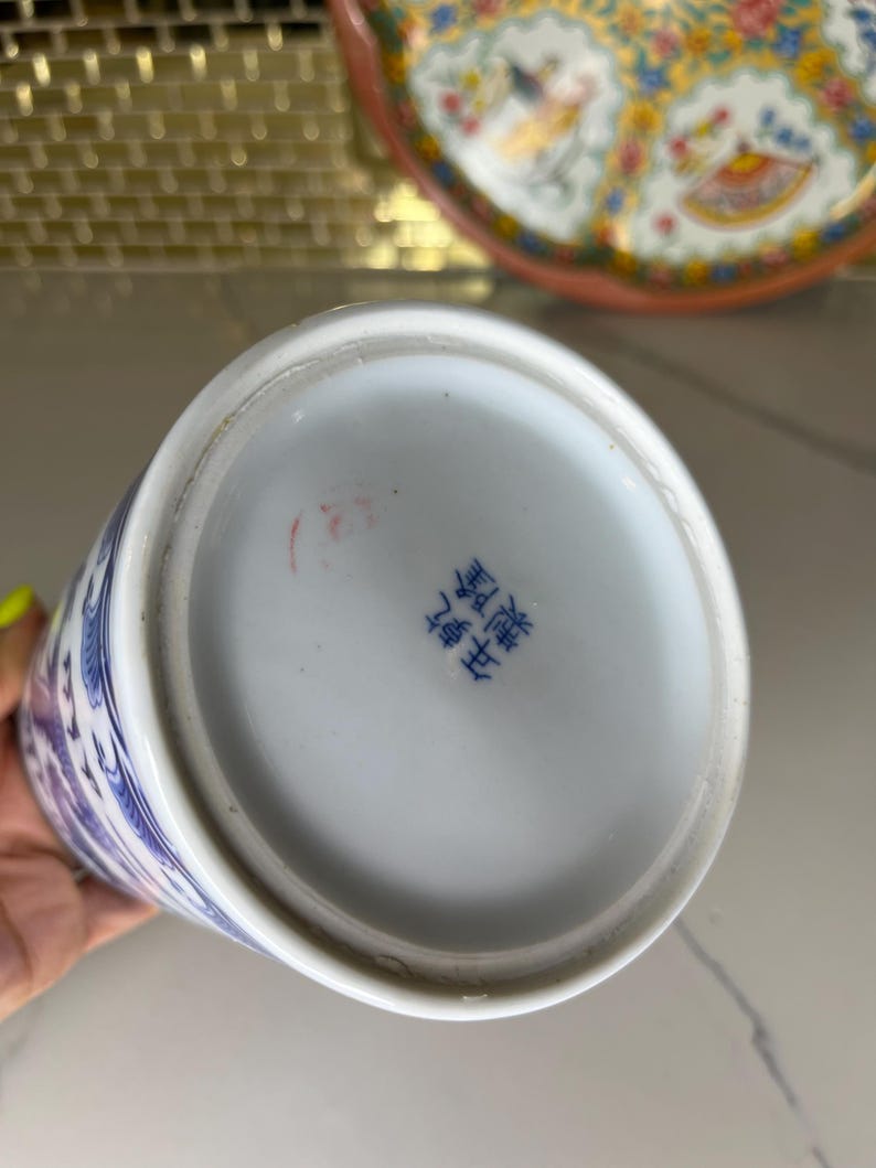 May include: A white and blue porcelain vase with a floral design. The bottom of the vase has blue Chinese characters. A decorative plate with a floral design is in the background.
