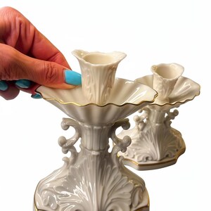 May include: Two white porcelain candle holders featuring a scalloped bobeche and a central socket. The base and rim are decorated with intricate embossed leaf patterns and finished with a thin gold trim along the edges of the base and the bobeche.