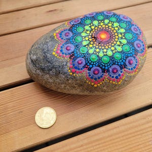 May include: A smooth, grey river stone decorated with a vibrant, multi-colored mandala pattern made of painted dots in shades of yellow, green, blue, purple, and pink. A gold-colored 50 cent coin is placed next to the stone for scale reference.