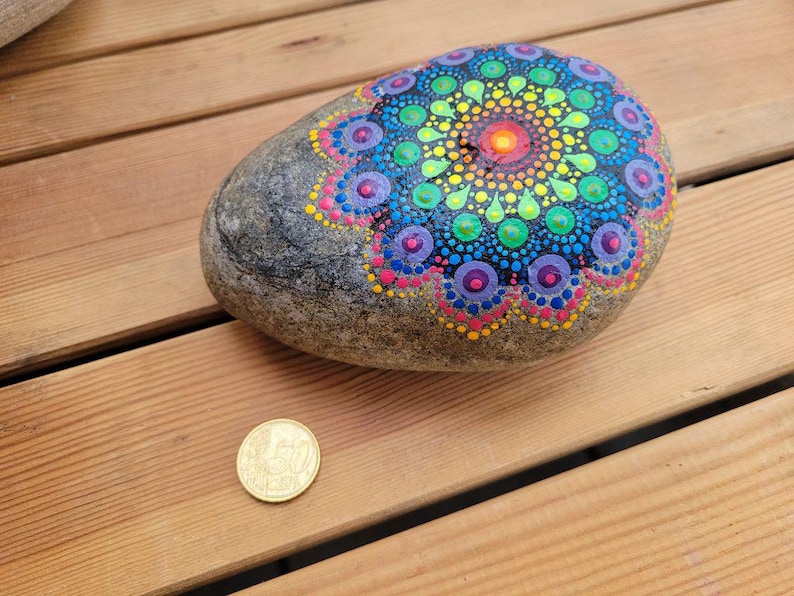 May include: A smooth, grey river stone decorated with a vibrant, multi-colored mandala pattern made of painted dots in shades of yellow, green, blue, purple, and pink. A gold-colored 50 cent coin is placed next to the stone for scale reference.