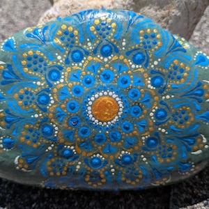 May include: A smooth, oval-shaped gray stone featuring a hand-painted mandala design. The artwork consists of concentric circles of dots and fine lines in vibrant shades of bright blue, metallic gold, and white, radiating from a central gold dot.