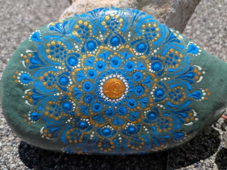 May include: A smooth, oval-shaped gray stone featuring a hand-painted mandala design. The artwork consists of concentric circles of dots and fine lines in vibrant shades of bright blue, metallic gold, and white, radiating from a central gold dot.