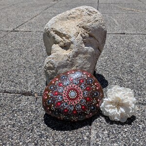 May include: A smooth, dark brown river stone decorated with a hand-painted mandala pattern in shades of red, white, and light blue. The intricate design features concentric circles of dots radiating from a central point, accompanied by a large rock.
