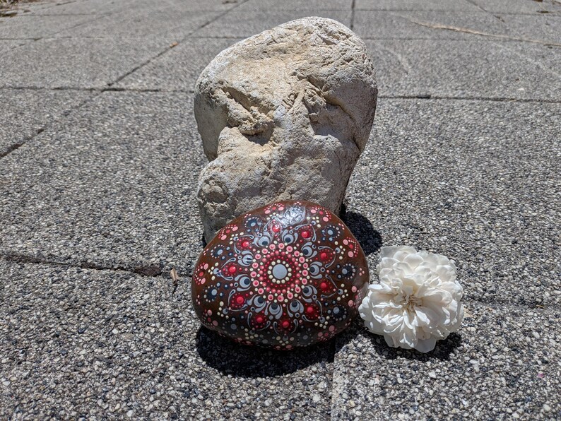 May include: A smooth, dark brown river stone decorated with a hand-painted mandala pattern in shades of red, white, and light blue. The intricate design features concentric circles of dots radiating from a central point, accompanied by a large rock.